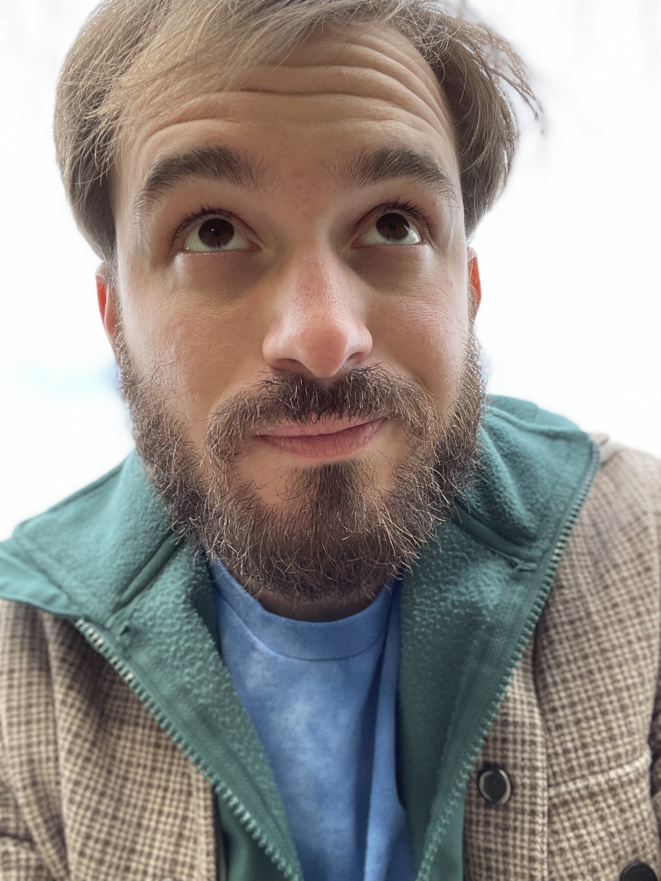 A self-portrait of Tyler Martinez wearing a teal hoodie layered under a tan jacket. He has medium-length brown hair and a full brown beard.  He is gazing past the camera at something in the distance.