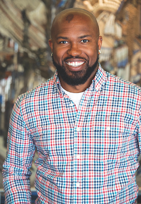 Pictured here is a photograph of Dr. Donnie Johnston Sackey. Dr. Johnson Sackey is smiling and looks directly at the camera with his arms at his sides. He wears earrings, and wears a plaid shirt with red, dark blue, and light blue stripes in the plaid pattern. Behind him is a brown multicolor patterned background.