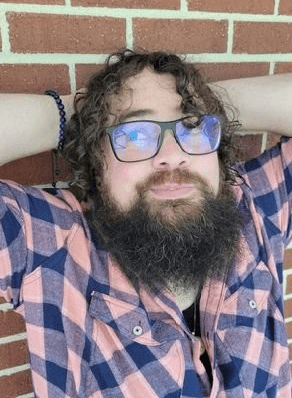 Pictured here is Ada Hubrig. They are seated with their arms casually raised behind their head. Ada wears black rectangular glasses, a black beaded bracelet, and is wearing a pink and dark blue plaid shirt. Behind them is a brick wall. Ada has brown curly hair and a brown beard.