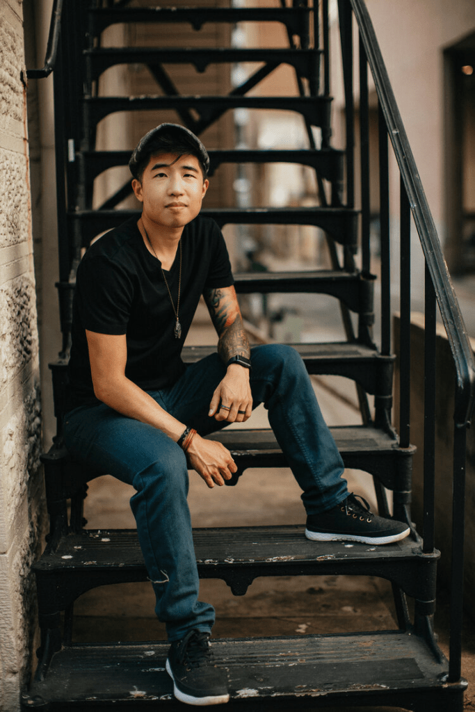 Pictured here is Dr. Vox Jo Hsu who is sitting on a black staircase that has chipped paint. They are wearing black sneakers, straight leg blue jeans, and a black tee shirt. They have short black hair, are wearing black and red bracelets, and have black and red tattoos on their left arm.