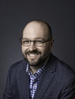 Pictured here is Benjamin Gatling, shown from the chest up. He wears a purple and white plaid collared shirt with a dark gray blazer over it. Behind him there is a dark gray background.