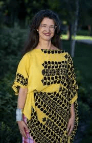 Pictured here is Libby Catchings who is shown smiling at the camera, pictured from the waist up. She wears a yellow and black floral patterned wrap shirt and her arms are at her sides. Behind her there are trees.
