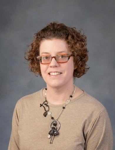 Pictured here is Sandra Faulkner. She sits in front of a gray background. She has short ear-length curly red hair and wears red and yellow glasses. She is smiling, and is wearing a black and brown necklace with a light brown shirt.