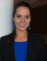 Pictured here is Anna Habib who is shown from the chest up. She wears a black blazer with a blue shirt and blue earrings. Her hair is brown and in a pony tail. Behind her there is a white and brown wall.