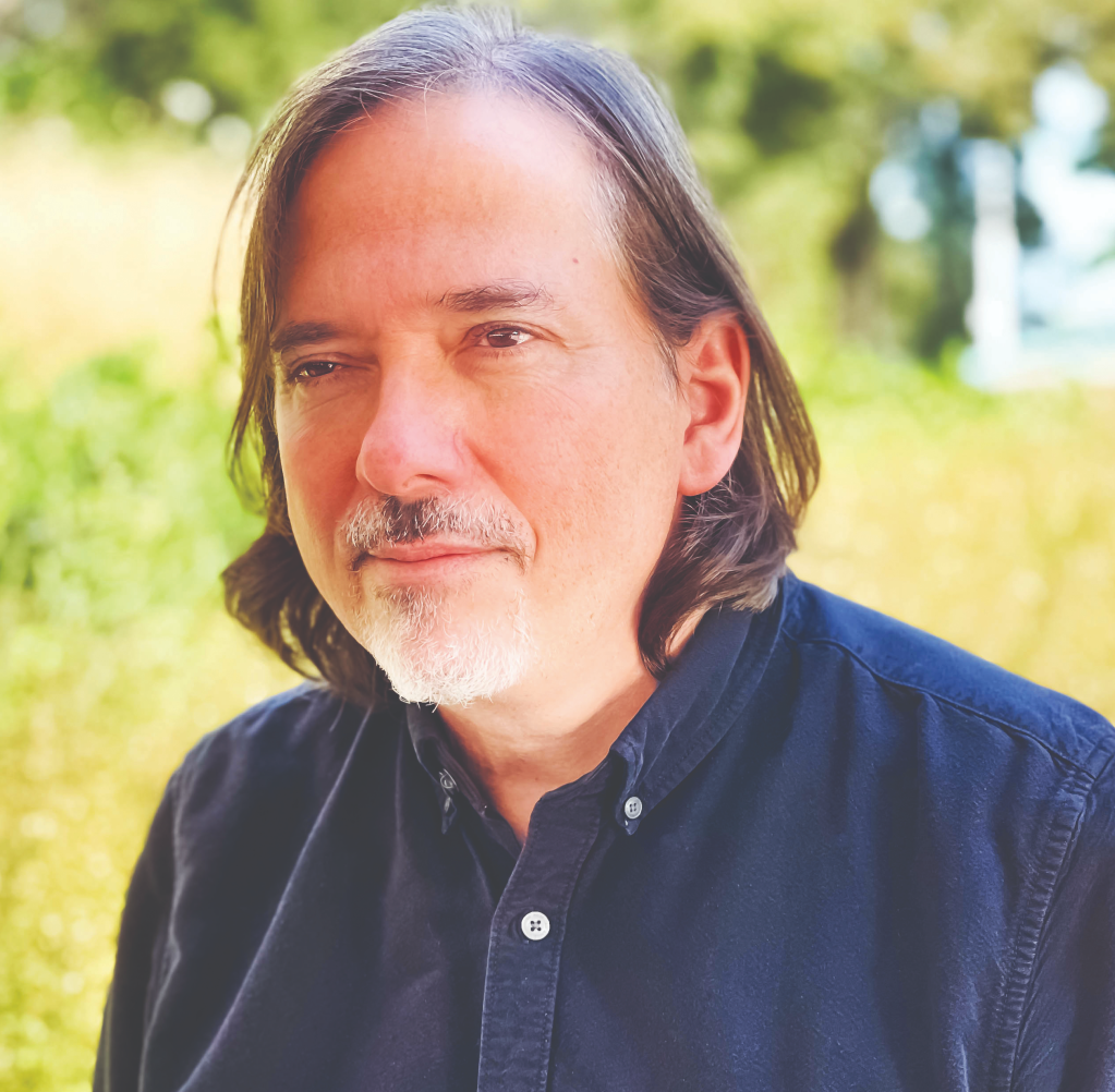 Patrick Berry is pictured looking off to the left. He wears a dark blue buttoned and collared shirt and has almost shoulder length brown hair. Behind him there is a fuzzy, unclear green background that might be trees and grass.